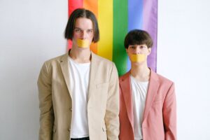 Two men with taped mouths stand against a rainbow flag, symbolizing LGBTQ silence and oppression.