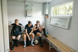 Female athletes in sports attire resting on a bench in a locker room with soccer gear.