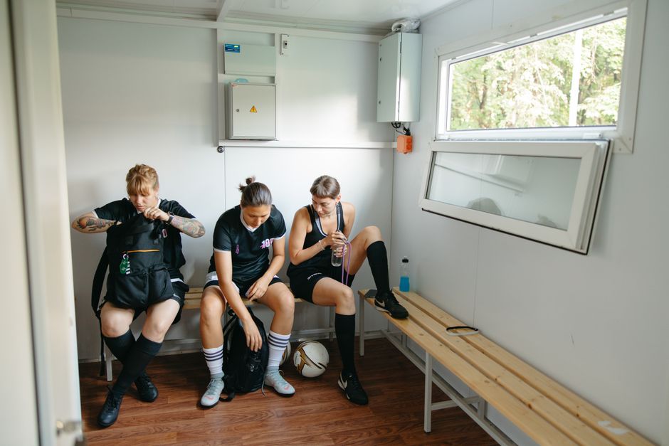 Female athletes in sports attire resting on a bench in a locker room with soccer gear.