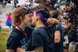Two women share a kiss at a vibrant outdoor pride festival, celebrating love and inclusivity.